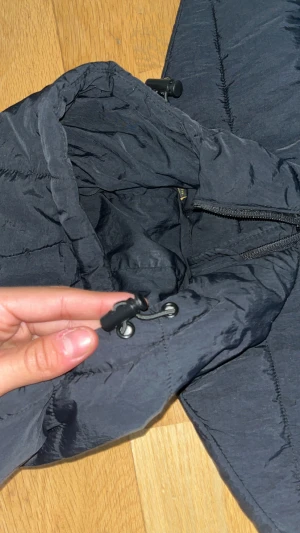 Svart pufferjacka från Lyle & Scott - Svart pufferjacka från Lyle & Scott med gul broderad logga på bröstet. Jackan har huva med snörning, dragkedja framtill och quiltad design. Perfekt för kalla dagar och har en clean, modern look som funkar till streetwear.