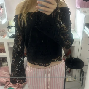 Svart offshoulder spetsblus - Säljer en svart blus med offshoulder-design och långa, vida ärmar. Blusen är helt i spets med transparenta partier och har en croppad passform. Spetsen ger en snygg och detaljerad look.
