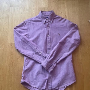 Lila slim fit skjorta från Ralph Lauren - Snygg lila skjorta från Ralph Lauren i slim fit-modell, stretchig oxfordväv i bomull. Klassisk krage, knappar framtill och broderad logga på bröstet. Perfekt för dig som gillar stilrena och ikoniska plagg med en modern passform.