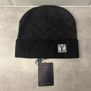 Svart Louis Vuitton mössa med logga - Snygg svart mössa från Louis Vuitton med diskret rutigt mönster och vikt kant. På framsidan sitter en vit patch med LV-loggan. Mössan är tillverkad i mjukt material och passar perfekt för kalla dagar.