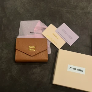 Brun plånbok från Miu Miu i skinn - Snygg brun plånbok från Miu Miu i äkta skinn med guldfärgad logga framtill. Plånboken har flera fack för kort och sedlar, samt en praktisk knappstängning. Kommer med original ask och certifikat. Perfekt storlek för att ha med sig överallt.