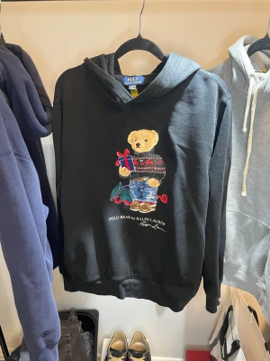 Svart Polo Bear hoodie från Ralph Lauren - Svart hoodie från Ralph Lauren med Polo Bear-tryck på framsidan där björnen håller en present. Tröjan har huva och ribbade muddar. Materialet är mjuk bomull och polyester, perfekt för dig som gillar ikoniska motiv och chill stil.