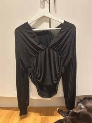 Svart omlott långärmad topp från NLY ONE - Säljer en svart omlott topp från NLY ONE i mjuk polyester. Toppen har lång ärm, v-ringad hals och snygga draperade detaljer framtill som ger en cool och modern look. Perfekt för dig som gillar att sticka ut med din stil.