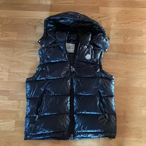 Svart dunväst från Moncler med huva - Svart glansig dunväst från Moncler med avtagbar huva och två dragkedjefickor framtill. Västen har quiltad design och klassisk Moncler-logga på bröstet. Perfekt för lager-på-lager och snygg till streetwear.