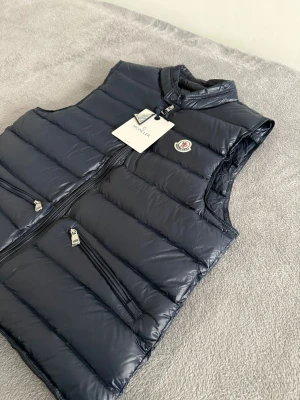 Moncler väst  - Snygg mörkblå dunväst från Moncler med quiltad design och hög krage. Västen har två snedställda fickor med dragkedja och ett klassiskt Moncler-märke på bröstet. Materialet är glansigt och känns lyxigt, perfekt för att lyfta din look.