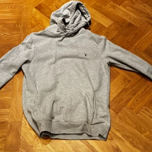 Grå hoodie från All Saints - Säljer en grå hoodie från All Saints med liten broderad logga på bröstet. Tröjan har huva med snörning, ribbade muddar och en klassisk loose passform. Perfekt för chill dagar och enkel att matcha med jeans eller joggers.