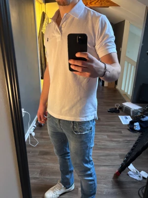 Vit pikétröja från Ralph Lauren - Snygg vit pikétröja från Ralph Lauren med klassisk krage och korta ärmar. Tröjan har den ikoniska broderade loggan i svart på bröstet.