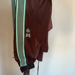 Needles x Girls Don't Cry bruna trackpants - Bruna trackpants från Needles x Girls Don't Cry med turkosa ränder längs sidorna, broderad fjäril och hjärta och text på låret. Byxorna har elastisk midja, dragkedjefickor och är gjorda i glansig polyester. Perfekt för en streetwear-look.