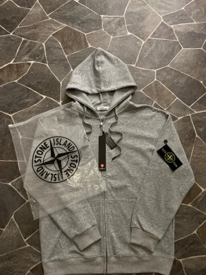 Grå hoodie från Stone Island - Snygg grå hoodie från Stone Island med klassisk logotyp-patch på vänster ärm. Tröjan har huva med snörning, dragkedja framtill och två fickor. Tillverkad i mjuk bomull för skön komfort. Perfekt för dig som gillar streetwear och stilrena detaljer.