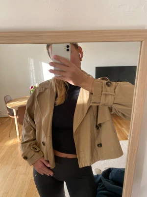 Beige croppad trenchcoat med knappar - Säljer en beige croppad trenchcoat med dubbelknäppning och stora mörka knappar. Jackan har bred krage, justerbara remmar vid ärmslut och är tillverkad i bomull. Perfekt för dig som gillar en chill och trendig look.Har en liten fläck vid krage, skriv privat så skickar jag bild.
