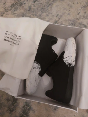 Maison Margiela svartvita sneakers - Maison Margiela sneakers med unik svart och vit färgblockad design. Skorna har snörning, platt sula och rund tå. Materialet är canvas och skinn, med en cool handmålad look på tån och sidorna. Märkets logga syns på sulan och dustbag ingår.KAN GÅ NED I PRIS!!