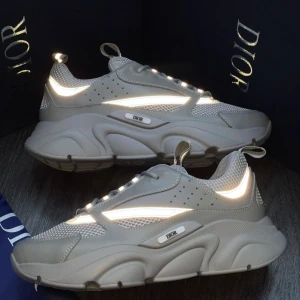 Grå sneakers från Dior - Säljer ett par grå  sneakers från Dior med reflekterande detaljer och meshpaneler. Skorna har en kraftig plattformssula, snörning och rund tå. Perfekt för dig som gillar exklusiv streetstyle och vill sticka ut med ett lyxigt märke.
