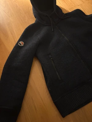 Mörkblå stickad hoodiejacka Superdry - Mörkblå hoodiejacka från Superdry med stickad utsida och mjuk fleece på insidan. Storlek XL på insidan av någon anledning men passar som M bra för 180-185 cm lång. Jackan har huva med snörning, två fickor med dragkedja och Superdry Expedition-märke på ärmen. Perfekt för kalla dagar och chill stil.