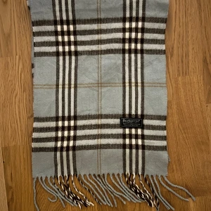 Burberry Halsduk  - Snygg rutig halsduk från Burberrys i 100% lammull, tillverkad i England. Färgerna går i ljusgrått, beige och brunt med klassiskt rutmönster och fransar i ändarna. Perfekt accessoar för att lyfta din outfit med lite brittisk stil.