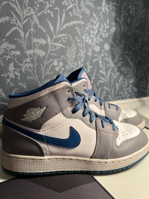 Nike Air Jordan 1 Mid grå/blå sneakers - Nike Air Jordan 1 Mid sneakers i grått och vitt skinn med blå detaljer och blå snörning. Klassisk Jordan-logga på sidan och tungan. Snygg kontrast mellan de olika färgblocken och den blå swooshen. Perfekta för dig som gillar streetstyle och ikoniska sneakers.