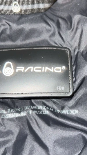 Svart pufferjacka från Sail Racing - Svart pufferjacka från Sail Racing med huva och dragkedja framtill. Jackan har två sidofickor med dragkedja och logga på bröstet och huvan. Materialet är glansigt och känns lätt men ändå isolerande, perfekt för kyligare dagar.