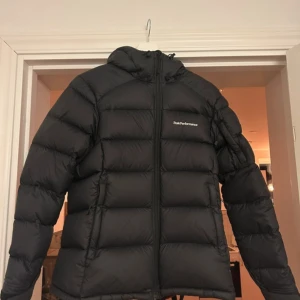 Svart dunjacka Peak Performance S - Svart dunjacka från Peak Performance med huva och dragkedja framtill. Jackan är quiltad och har en clean look med diskret logga på bröstet. Yttermaterialet är Pertex Quantum som är lätt och slitstarkt. Perfekt för kalla dagar. JätteFint skick förutom att den har en liten lagning längst ner på ryggen som du ser på bilden men inget som syns riktigt! Pris kan diskuteras 