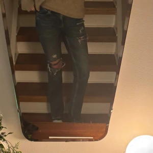 Blå slitna jeans  - Säljer ett par blå jeans med snygga slitningar och hål vid knäna. Modellen är Bootcut och klassisk femficksdesign. Jeansen har en mörk tvätt och är tillverkade i jeansmaterial med dragkedja och knapp framtill.