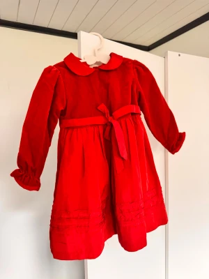 Röd sammetsklänning med rosett för barn - Supersöt röd sammetsklänning med rund krage, långa puffärmar och dekorativ rosett i midjan. Klänningen har knappar baktill och fina volangdetaljer vid ärmslut och nederkant. Perfekt för små barn storlek 80cm