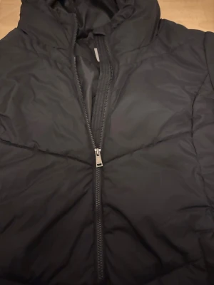 Svart lång pufferjacka Cellbes - Svart pufferjacka från Cellbes med huva och långa ärmar. Jackan har snygga diagonala quiltade sömmar, hel dragkedja framtill och praktiska fickor med knapp. Perfekt för kalla dagar och har en clean, stilren look.