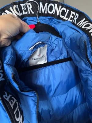 Blå Moncler pufferjacka med huva - Säljer en blå pufferjacka från Moncler med stor huva och breda svarta band med vit Moncler-logga. Jackan har dragkedjor på sidorna och klassisk Moncler-patch på ärmen. Passar för ca 10-12år Strl ca 152cm