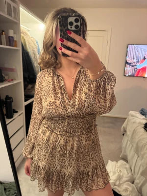 Leopardmönstrad kortklänning med volang - Säljer en superfin kortklänning med leopardmönster i beige och bruna toner. Klänningen har v-ringad hals, långärmade puffärmar och smock i midjan för en snygg siluett. Volangdetaljer längst ner ger en extra touch. Perfekt för dig som gillar trendiga och djuriska prints.