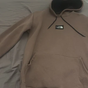 Brun hoodie från The North Face - Säljer en brun hoodie från The North Face med svart snörning och logga på bröstet. Tröjan har en stor huva, magficka och ribbade muddar vid ärmslut och nederkant. Perfekt för chill dagar och streetwear-stil.