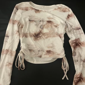 Långärmad topp med knyt och mönster - Säljer en långärmad topp i ljusa toner med abstrakt mönster i beige och brunt. Toppen har snygga knytband i sidorna som ger en cool ruche-effekt och croppad passform. Materialet känns mjukt och stretchigt, perfekt till jeans eller kjol.
