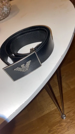Svart skärp från Emporio Armani äkta läder - Snyggt svart bälte från Emporio Armani i äkta skinn med en stilren, rektangulär metallspänne och den klassiska Armani-loggan i silver. Perfekt accessoar för att lyfta din outfit med en exklusiv touch. Tillverkat i Italien. Helt äkta i läder storlek 75/30. 1000kr nytt
