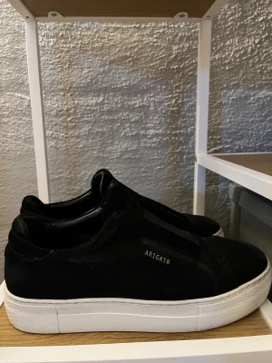 Svarta sneakers från Axel Arigato - Stilrena svarta sneakers från Axel Arigato i mocka med vit platt sula. Diskret guldtext på sidan och klassisk rund tå.  Inte alls använda mycket så är i ett väldigt bra skick! Perfekt för både vardags och om man vill klä upp sig. Pris kan diskuteras!