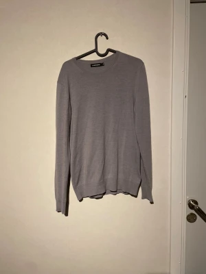 Grå stickad tröja från J.Lindeberg - Snygg grå stickad tröja från J.Lindeberg med diskret logga på bröstet. Tröjan har rund hals, ribbade muddar och lång ärm. Perfekt för dig som gillar stilrena och enkla plagg med en modern touch.