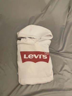 Vit hoodie från Levi's med röd logga storlek 140 - Säljer en vit hoodie från Levi's i storlek 140 cm. Tröjan har en stor röd Levi's-logga på bröstet och klassisk huva. Mjuk insida och ribbade muddar vid ärmslut och nederkant. Perfekt för dig som gillar streetwear och vill ha en clean look. Den lilla svarta pricken går ej bort men annars jätte bra sick pricken syns knappt och ät nån millientmemeter 