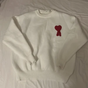 Vit stickad tröja från Ami med rött hjärta - Vit stickad tröja från Ami med ett stort rött hjärta och A broderat på bröstet. Tröjan har rund hals, ribbade muddar och nederkant samt långärmad passform. Perfekt för dig som gillar clean stil med en twist. Aldrig andvände innan Storlek S men passar också M 