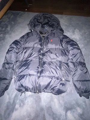  Polo Ralph Lauren jacka - Svart pufferjacka från Polo Ralph Lauren med huva och dragkedja framtill. Jackan har två sidofickor med dragkedja och ett rött Polo-logomärke broderat på bröstet. Materialet är glansigt och känns riktigt chill för kalla dagar.