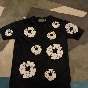 Svart t-shirt med blommigt tryck Denim Tears - Svart t-shirt från Denim Tears med vita blommiga tryck över hela plagget. T-shirten har rund hals och korta ärmar. Materialet känns mjukt och lätt, perfekt för dig som gillar streetwear med unik design.