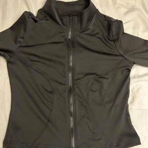 Svart sportig vindjacka med dragkedja - Svart, figursydd vindjacka i syntetmaterial med hel dragkedja framtill och hög krage. Jackan har långa ärmar och stilrena sömdetaljer som ger en sportig vibe. Perfekt för dig som gillar en clean och enkel look.