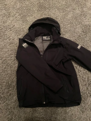 Svart vindjacka Helly Hansen med huva - Svart vindjacka från Helly Hansen med vit HH-logga på axel och ärm. Jackan är i bra skicka några tecken på användning! Jackan är i storlek S/M