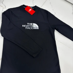 Svart långärmad tröja The North Face - Svart långärmad tröja från The North Face med klassisk logga i silver på bröstet. Tröjan har rund hals och är tillverkad i mjuk bomull som känns skön mot huden. Perfekt för dig som gillar stilrena och sportiga plagg med tydlig branding.