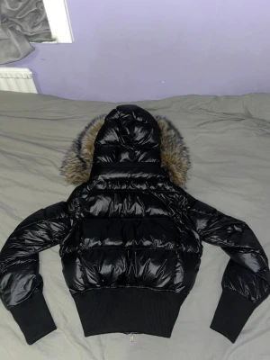 Svart croppad pufferjacka med fuskpäls - Säljer en svart, croppad pufferjacka med glansig finish och stor huva med fluffig fuskpäls. Jackan har dragkedja framtill och sitter tajt i midjan för en snygg siluett. Perfekt för dig som vill ha en trendig vinterlook.