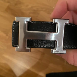 Svart skärp med H-spänne - Säljer ett svart bälte i skinn med det ikoniska H-spännet i silverfärgad metall. Bältet har en stilren och klassisk design med vita sömmar längs kanterna. Perfekt för att lyfta din outfit med en lyxig touch.