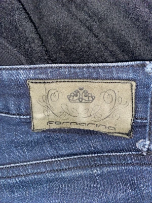 Mörkblå jeans från Fornarina - Snygga mörkblå jeans från Fornarina med klassisk femficksdesign och coola slitningar. Jeansen har en röd detalj i midjan och en broderad logga på bakfickan. Passformen är rak och materialet är stretchigt denim för extra komfort.