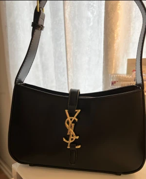 Svart axelväska från Saint Laurent - Snygg svart axelväska från Saint Laurent i slätt skinn med guldiga YSL-detaljer framtill. Väskan har en stilren, rektangulär form och justerbar axelrem med guldfärgat spänne. Dragkedja upptill och elegant design som verkligen sticker ut.