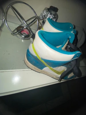 Nike Dunk High grön och vit sneakers - Nike Dunk High sneakers i blått, vitt och med en limegrön swoosh. Skorna har svart snörning, perforerad tå och högt skaft. Ovandelen är i skinn och textil, med platt sula och rund tå. Perfekt för dig som gillar färgstarka och klassiska sneakers.