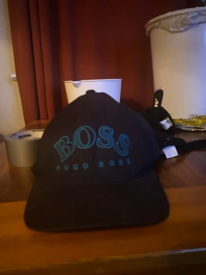 Svart keps från Hugo Boss - Snygg svart keps från Hugo Boss med tydlig BOSS-logga i blått framtill. Klassisk böjd skärm och justerbar baksida. Perfekt accessoar för en stilren och sportig look.