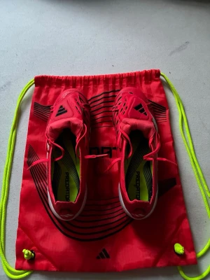 Adidas Predator röda fotbollsskor - Snygga röda Adidas Predator fotbollsskor med vita och svarta detaljer. Skorna har låg profil, snörning och en ovandel i syntetmaterial för bra bollkontroll. Kommer med matchande röd gympåse med neongula snören. Perfekta för dig som vill sticka ut på planen.