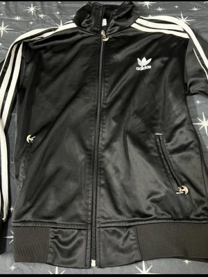 Svart Adidas Originals track jacket - Klassisk svart Adidas Originals track jacket med vita ränder längs ärmarna och dragkedja framtill. Tröjan har ståkrage, två fickor med dragkedja och ribbade muddar. Tillverkad i glansigt syntetmaterial och pryds av Adidas logga på bröstet.