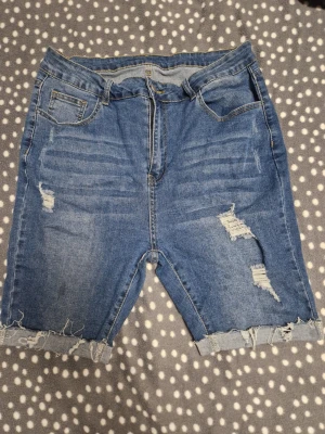 Blå jeansshorts med slitningar - Säljer ett par blå jeansshorts med råa, fransiga benslut och slitna detaljer framtill. Klassisk femficksmodell med dragkedja och knapp. Perfekta för sommaren och ger en avslappnad streetstyle-vibe.