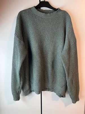 Grön oversized stickad tröja - En oversized stickad tröja i grågrönt med grov ribbstickning och rund hals. Tröjan har långa ärmar och avslappnad passform, perfekt för lager-på-lager. Materialet känns mjukt och bekvämt.