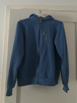 Blå hoodie från Sail Racing - Blå hoodie med dragkedja från Sail Racing. Tröjan har huva, bröstficka med dragkedja och logga tryckt på bröstet. Perfekt för en avslappnad stil och enkel att matcha med jeans eller joggers.