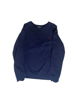Mörkblå sweatshirt från Polo Ralph Lauren - Mörkblå sweatshirt från Polo Ralph Lauren med rund halsringning och klassisk röd broderad logga på bröstet. Tröjan har långa ärmar och ribbade muddar vid ärmslut och nederkant. Perfekt för en avslappnad och stilren look.